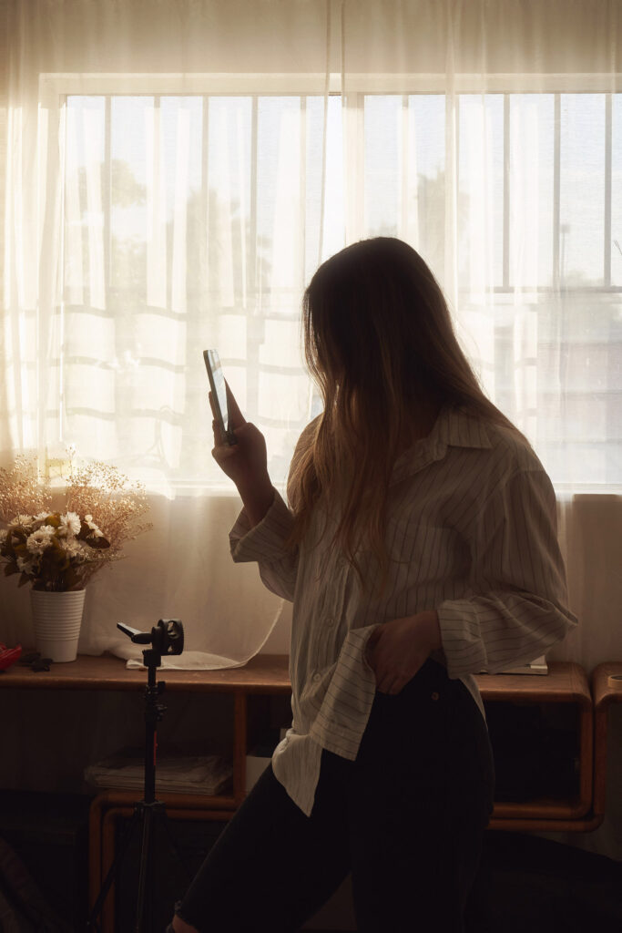Gaby and Me a sunlit room with a person in a striped shirt holding a phone a table with a vase of flowers and a tripod stand.